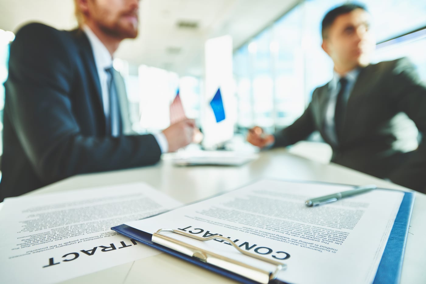 Contratti cartacei su un tavolo riunioni; sullo sfondo, sfocati, due professionisti in giacca